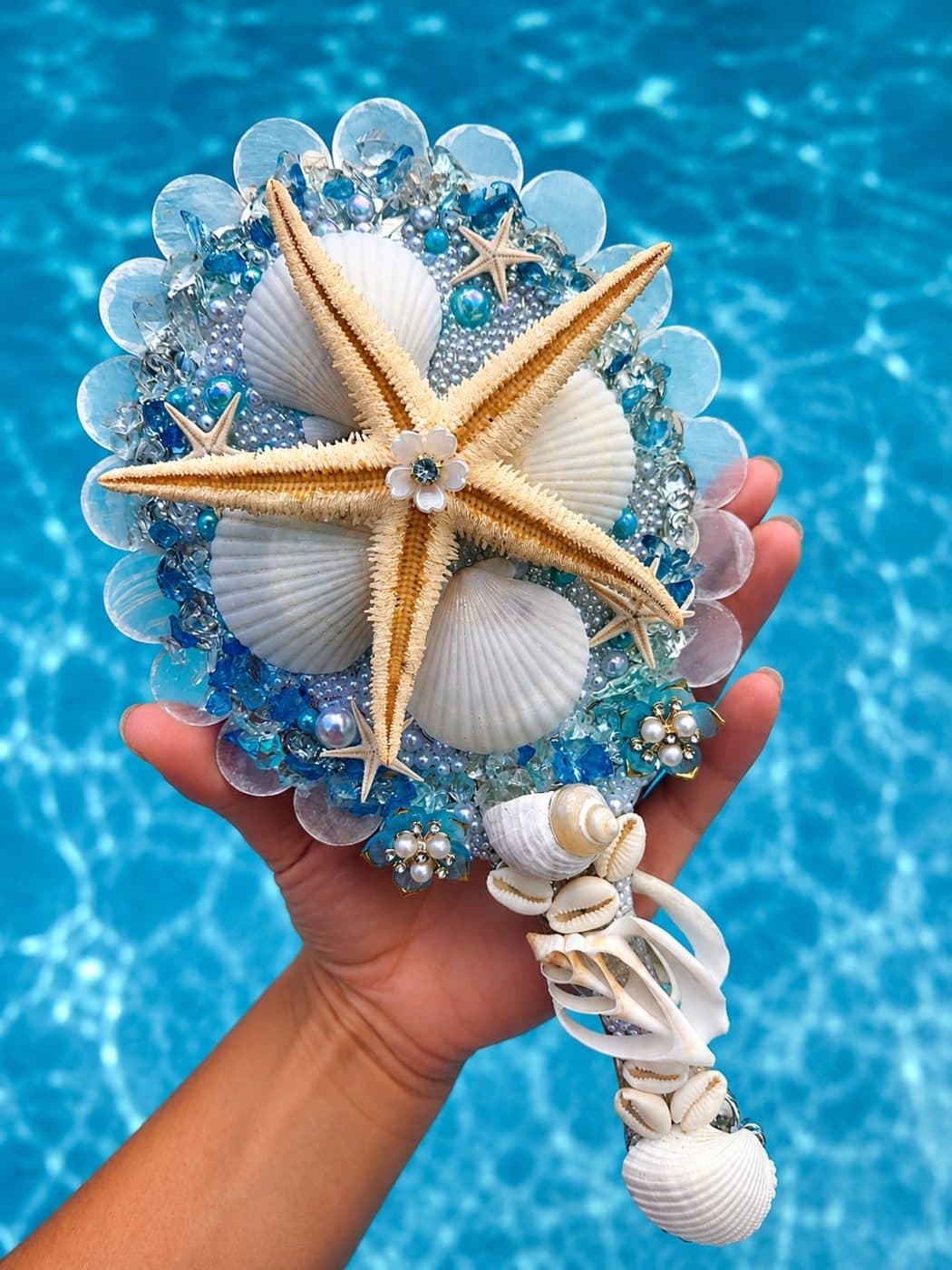 Espelho Reflexo das Marés com grande estrela-do-mar, conchas e pedrarias azuis, segurado diante de uma piscina cristalina