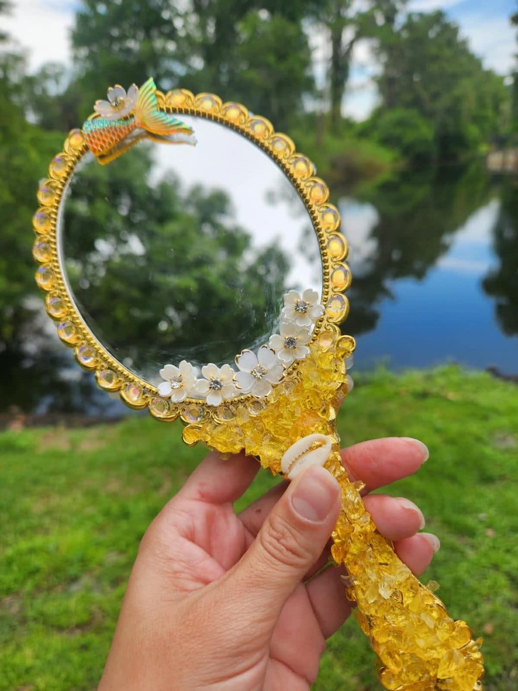 Espelho Reflexo de Areia com moldura de crushed glass dourado, flores brancas e miniatura de sereia, diante de um lago